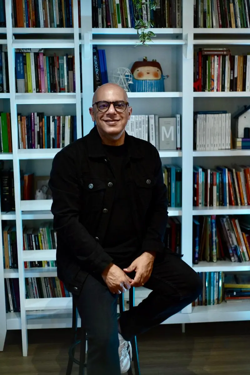 Flávio Azeredo de jaqueta preta e óculos, sentado num banco alto diante da estante de livros
