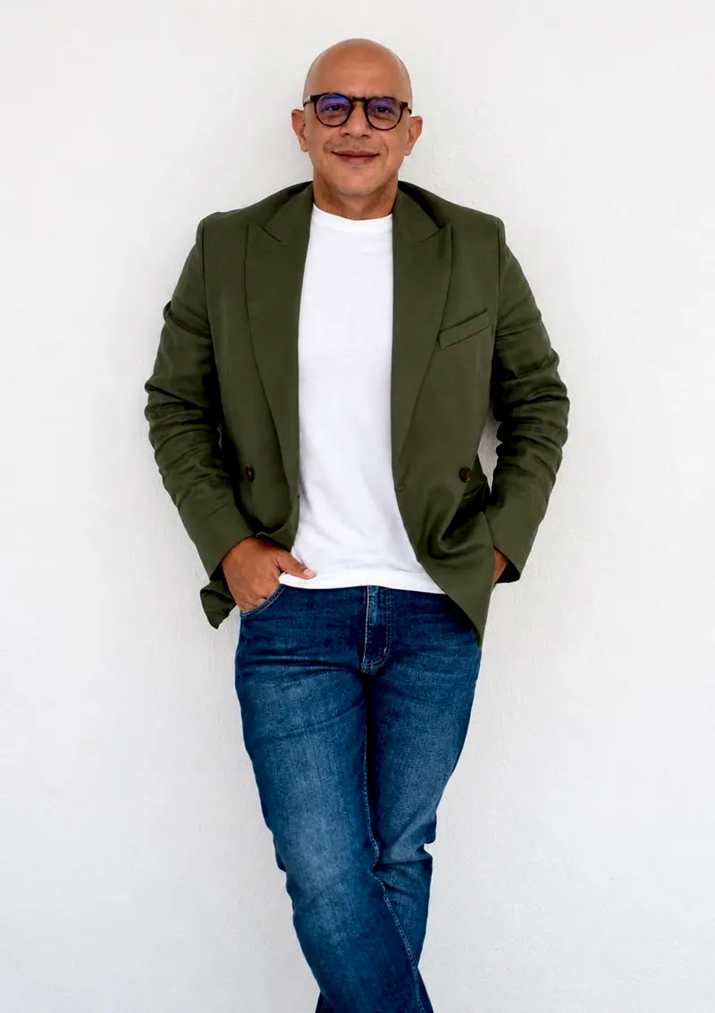 Flávio Azeredo de blazer verde, camiseta branca e óculos, encostado numa parede clara